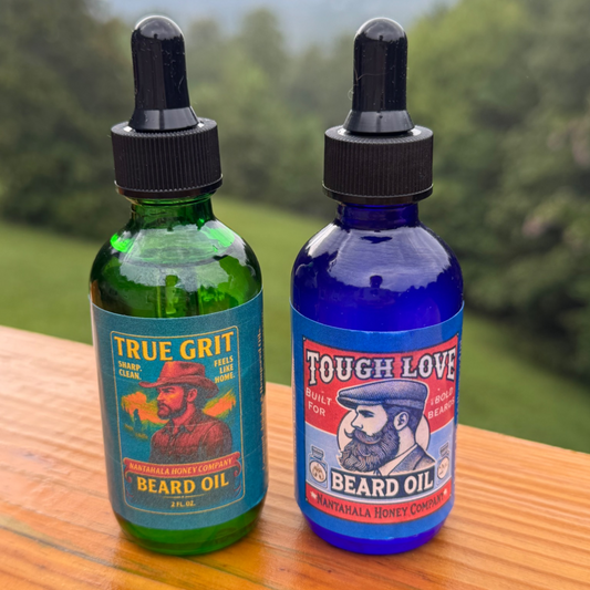 wo 2oz beard oil bottles from Nantahala Honey Co. on a wooden table with a mountain backdrop — True Grit in green glass and Tough Love in cobalt blue."